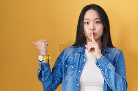 Téléchargez les photos : Jeune femme asiatique debout sur fond jaune demandant à être calme avec le doigt sur les lèvres pointant avec la main sur le côté. silence et concept secret. - en image libre de droit