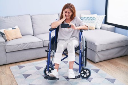 Téléchargez les photos : Middle age woman watching video on smartphone sitting on wheelchair at home - en image libre de droit