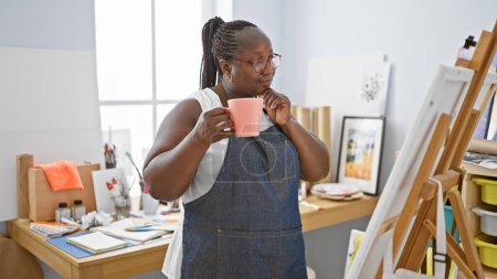 Téléchargez les photos : Femme artiste afro-américaine, debout avec une tasse de café, regardant son dessin dans son atelier d'art. - en image libre de droit