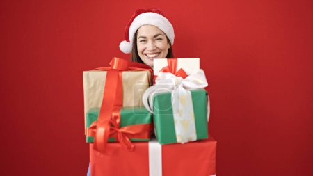 Téléchargez les photos : Jeune femme caucasienne portant chapeau de Noël tenant des cadeaux sur fond rouge isolé - en image libre de droit