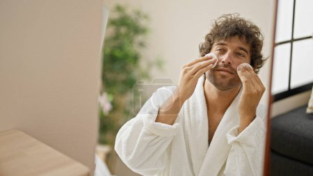 Téléchargez les photos : Jeune homme hispanique portant peignoir de nettoyage visage avec des tampons de coton regardant sur miroir à la maison - en image libre de droit