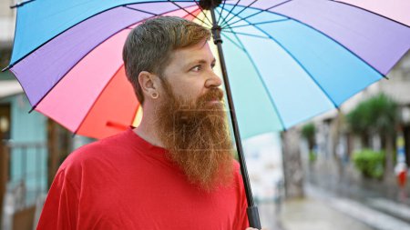 Photo for Cool redhead guy looking off to the side, rocking a serious expression while holding his umbrella on a sunny urban street - Royalty Free Image