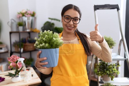 Foto de Mujer árabe joven que trabaja en floristería sosteniendo maceta sonriendo feliz y positivo, pulgar hacia arriba haciendo excelente y signo de aprobación - Imagen libre de derechos