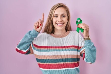 Photo for Young blonde woman holding support green ribbon surprised with an idea or question pointing finger with happy face, number one - Royalty Free Image