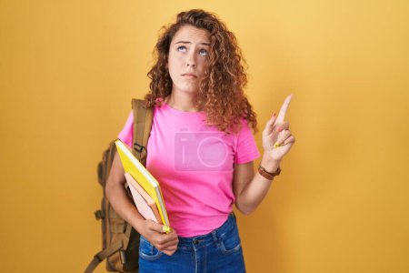 Foto de Young caucasian woman wearing student backpack and holding books pointing up looking sad and upset, indicating direction with fingers, unhappy and depressed. - Imagen libre de derechos