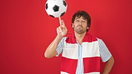 Foto de Joven hombre hispano apoyando equipo de fútbol jugando con pelota sobre fondo rojo aislado - Imagen libre de derechos