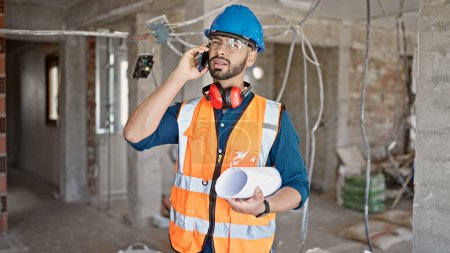 Téléchargez les photos : Jeune homme hispanique constructeur debout avec des plans parlant au téléphone sur le chantier de construction - en image libre de droit