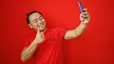 Téléchargez les photos : Jeune chinois insolent profitant d'un moment de selfie amusant sur un mur rouge isolé, donnant un geste confiant pouce vers le haut avec son smartphone. - en image libre de droit