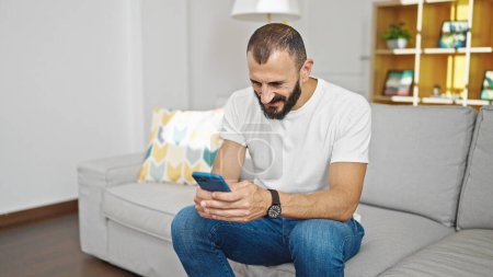 Téléchargez les photos : Jeune homme hispanique utilisant smartphone assis sur le canapé à la maison - en image libre de droit