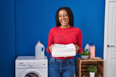 Photo for Young african american with braids holding clean laundry winking looking at the camera with sexy expression, cheerful and happy face. - Royalty Free Image