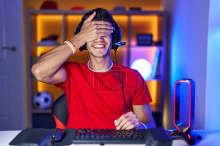 Foto de Joven hispano jugando videojuegos sonriendo y riendo con la mano en la cara cubriendo los ojos para sorpresa. concepto ciego. - Imagen libre de derechos