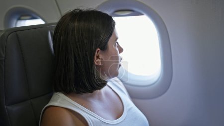 Foto de Joven hermosa mujer hispana sonriendo sentada mirando por la ventana en el avión - Imagen libre de derechos