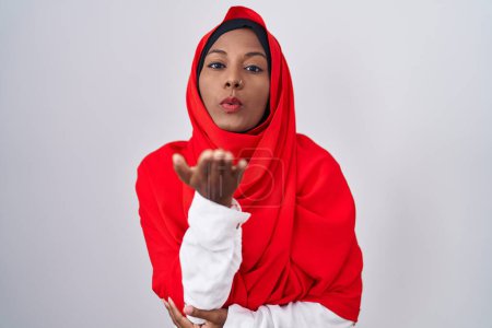 Foto de Mujer árabe joven vistiendo bufanda hijab islámica tradicional mirando a la cámara soplando un beso con la mano en el aire siendo encantador y sexy. expresión de amor. - Imagen libre de derechos
