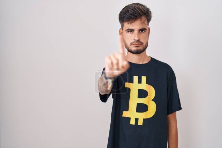 Téléchargez les photos : Jeune homme hispanique avec des tatouages portant Bitcoin t-shirt pointant avec le doigt vers le haut et expression en colère, ne montrant aucun geste - en image libre de droit