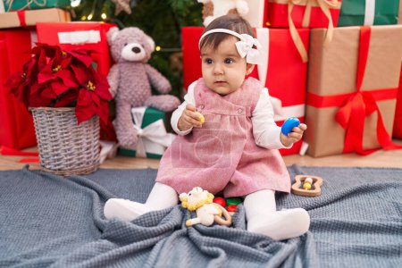 Photo for Adorable hispanic baby holding hoops toy sitting on floor by christmas tree at home - Royalty Free Image