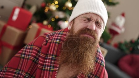 Photo for Young redhead man sitting on floor by christmas tree coughing at home - Royalty Free Image