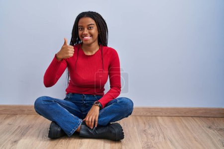 Téléchargez les photos : Jeune afro-américain avec des tresses assises sur le sol à la maison faisant des pouces heureux geste avec la main. approuvant l'expression regardant la caméra montrant le succès. - en image libre de droit