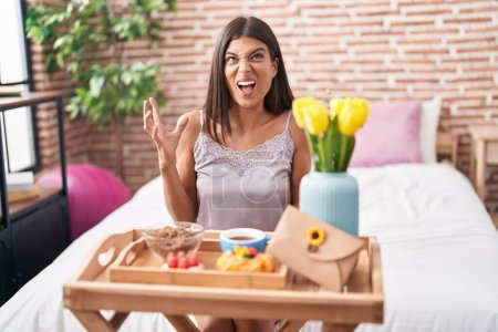 Photo for Brunette young woman eating breakfast sitting on the bed crazy and mad shouting and yelling with aggressive expression and arms raised. frustration concept. - Royalty Free Image