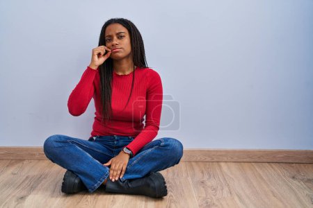 Téléchargez les photos : Jeune afro-américain avec des tresses assises sur le sol à la maison bouche et les lèvres fermées comme zip avec les doigts. secret et silencieux, tabou parlant - en image libre de droit
