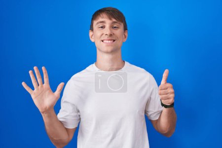 Foto de Caucásico hombre rubio de pie sobre fondo azul mostrando y señalando hacia arriba con los dedos número seis mientras sonríe confiado y feliz. - Imagen libre de derechos