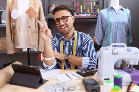 Photo for Hispanic man with beard dressmaker designer working at atelier smiling with happy face winking at the camera doing victory sign with fingers. number two. - Royalty Free Image