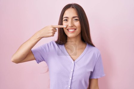 Téléchargez les photos : Jeune femme hispanique avec les cheveux longs debout sur fond rose pointant avec le doigt de la main vers le visage et le nez, souriant gai. concept de beauté - en image libre de droit