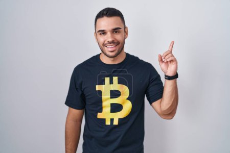 Téléchargez les photos : Jeune homme hispanique portant Bitcoin t-shirt avec un grand sourire sur le visage, pointant avec le doigt de la main sur le côté en regardant la caméra. - en image libre de droit