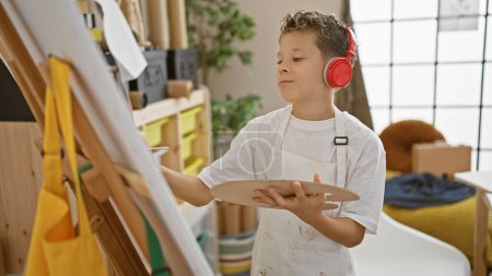 Foto de Adorable artista niño rubio, absorto con confianza en el dibujo, escuchando música en un estudio de arte, demostrando creatividad y concentración entre pinceles y pintura sobre lienzo. - Imagen libre de derechos