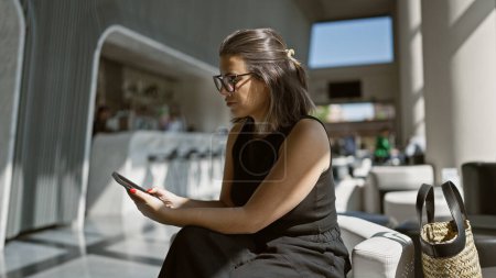 Téléchargez les photos : Belle femme hispanique à lunettes, occupée à envoyer des SMS sur son smartphone tout en se prélassant dans un hall d'hôtel moderne - en image libre de droit