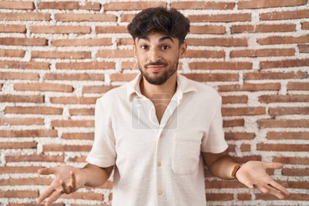 Photo for Arab man with beard standing over bricks wall background clueless and confused with open arms, no idea concept. - Royalty Free Image