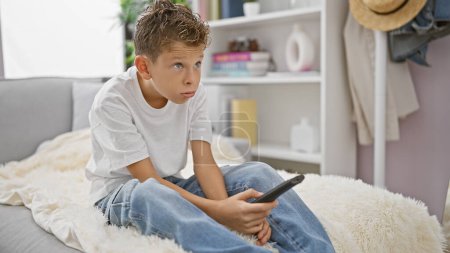 Photo for Adorable blond kid wearing a bored expression, comfortably hustling on the sofa at home, seeming serious while relaxing and watching tv in the living room - Royalty Free Image