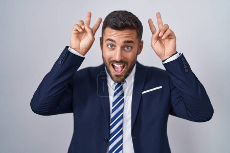 Foto de Hombre hispano guapo usando traje y corbata posando divertido y loco con los dedos en la cabeza como orejas de conejo, sonriendo alegre - Imagen libre de derechos