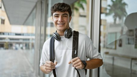 Téléchargez les photos : Joyeux jeune étudiant hispanique mâle debout à l'extérieur profitant de la vie sur le campus, portant avec confiance écouteurs et sac à dos, rayonnant d'énergie positive et de joie à l'université. - en image libre de droit