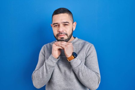 Téléchargez les photos : Homme hispanique debout sur fond bleu riant nerveux et excité avec les mains sur le menton regardant vers le côté - en image libre de droit