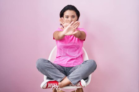 Téléchargez les photos : Jeune femme hispanique assise sur une chaise sur fond rose expression de rejet croisement des bras et des paumes faisant signe négatif, visage en colère - en image libre de droit