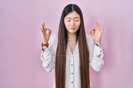 Photo for Chinese young woman standing over pink background relaxed and smiling with eyes closed doing meditation gesture with fingers. yoga concept. - Royalty Free Image
