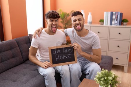 Photo for Homosexual gay couple holding blackboard with first home text sticking tongue out happy with funny expression. - Royalty Free Image
