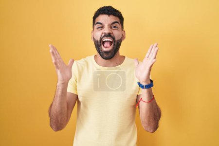 Téléchargez les photos : Homme hispanique avec barbe debout sur fond jaune célébrant fou et étonné pour le succès avec les bras levés et les yeux ouverts hurlant excité. concept gagnant - en image libre de droit