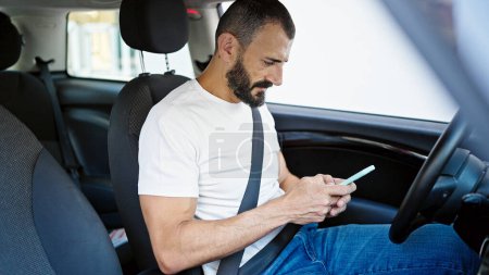 Foto de Young hispanic man using smartphone sitting on car at street - Imagen libre de derechos