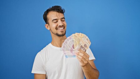 Foto de Joven hombre hispano sonriendo confiado sosteniendo pesos mexicanos sobre aislado fondo azul - Imagen libre de derechos