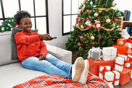 Foto de Mujer afroamericana viendo la televisión sentada en el sofá por el árbol de Navidad en casa - Imagen libre de derechos