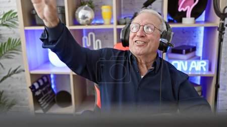 Téléchargez les photos : Streamer homme âgé énergique, avec cheveux gris et casque, dansant avec enthousiasme tout en streaming un jeu dans une salle de jeux faiblement éclairée - en image libre de droit