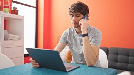Photo for Young hispanic man talking on smartphone using laptop at dinning room - Royalty Free Image