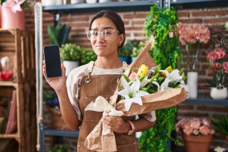 Photo for Young hispanic woman working at florist shop showing smartphone screen clueless and confused expression. doubt concept. - Royalty Free Image