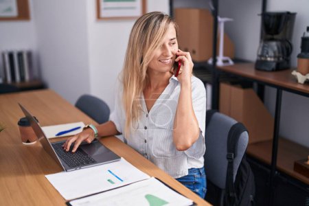 Téléchargez les photos : Jeune femme blonde travailleuse d'affaires utilisant un ordinateur portable parlant sur un smartphone au bureau - en image libre de droit