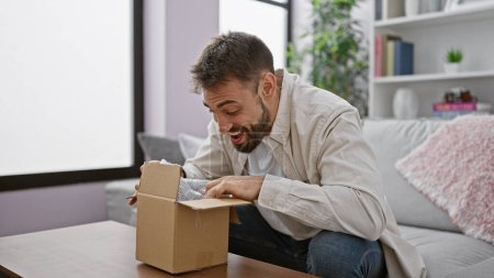 Photo for Young, confident hispanic man with a handsome beard happily unpacking a cardboard delivery box while sitting on a sofa at home, enjoying the indoor interior of his apartment living room - Royalty Free Image