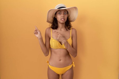 Téléchargez les photos : Jeune femme hispanique portant un bikini et un chapeau d'été pointant de côté inquiet et nerveux avec les deux mains, préoccupé et expression surprise - en image libre de droit