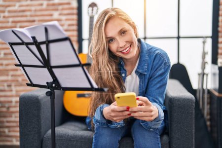 Photo for Young blonde woman musician using smartphone sitting on sofa at music studio - Royalty Free Image