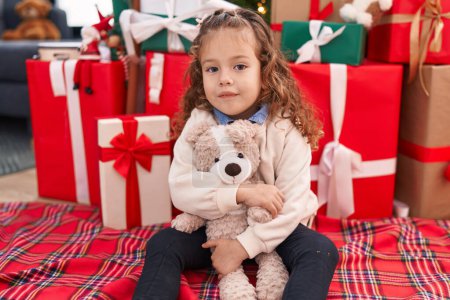 Foto de Adorable rubia niño abrazando osito de peluche sentado en el suelo por el árbol de Navidad en casa - Imagen libre de derechos