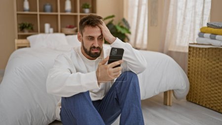 Téléchargez les photos : Stressé jeune homme hispanique assis seul dans une chambre à coucher, textant anxieusement sur son téléphone, luttant contre les maux de tête du matin et s'inquiéter - en image libre de droit